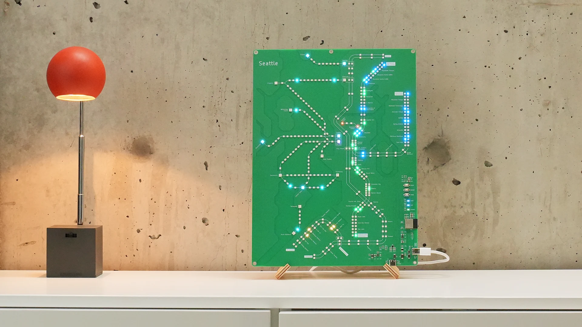 Seattle Transit Live illuminated in its birch plywood stand on a cabinet beside a red lamp